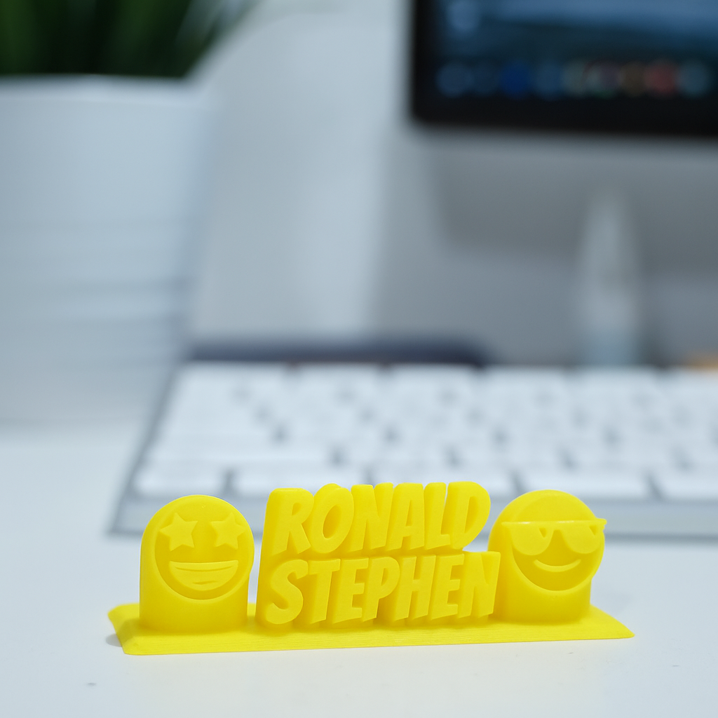 Yellow 3D-printed nameplate with 'RONALD STEPHEN' on a desk with a blurred keyboard and screen in the background.